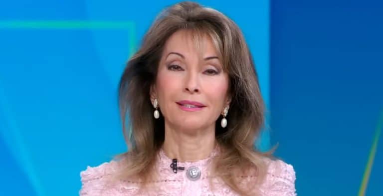 A woman with brown hair wearing a pink shirt with a blue background behind her.